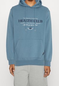 Felpa con cappuccio blu in tessuto morbido, con tasca a marsupio frontale e coulisse. Grafica testuale con la scritta “HEALTH CLUB” in colori misti.