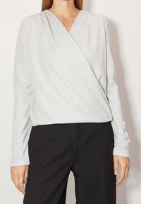 Top s dlouhým rukávem ve stylu zavinovacího topu z třpytivé stříbrné látky. Obsahuje výstřih do V, jemnou texturu a je kombinován s černými kalhotami.