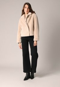 Chaqueta de piel sintética beige con cuello de muesca, cierre de doble botonadura y bolsillos laterales. Llevada sobre pantalones anchos negros y botas de tacón.