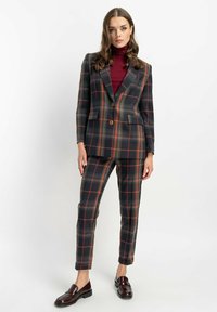 Kraciasty garnitur z dopasowaną marynarką i zwężanymi spodniami w ciemnozielonym, pomarańczowym i szarym kolorze. Gładka tkanina z dwoma guzikami i kieszeniami bocznymi. Brązowe loafersy.