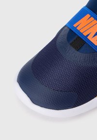 Zbliżenie na czubek granatowego sneakersa z teksturowanym syntetycznym materiałem, białą podeszwą i niebieską taśmą z pomarańczowym logo Nike.