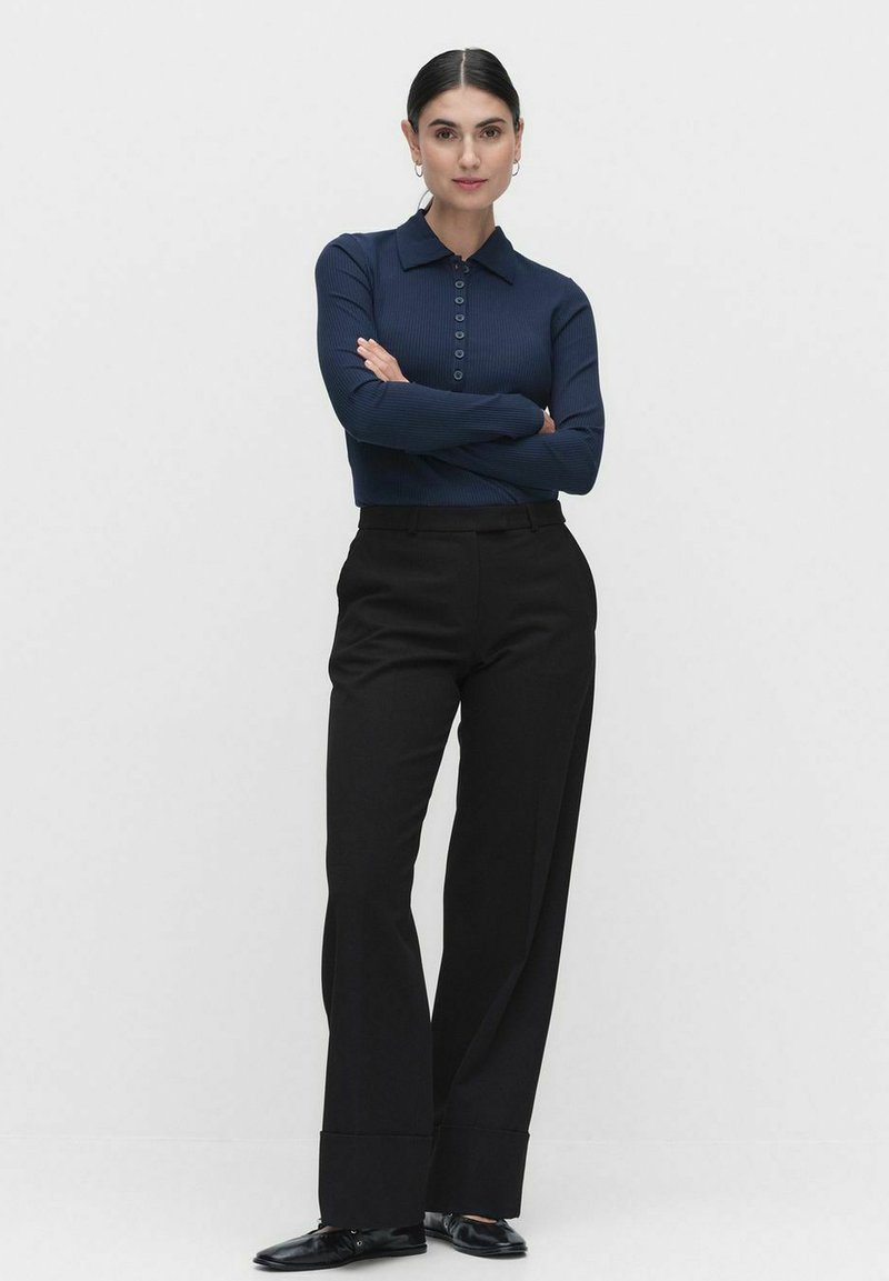 Tummansininen ribattu pitkähihainen polo paita, jossa on kaulus ja nappilista, yhdistettynä korkea vyötäröisiä mustia housuja ja mustia kenkiä.