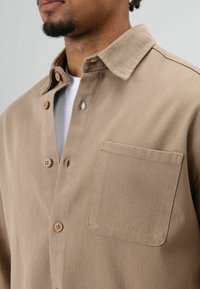 Homme portant une chemise beige à boutons par-dessus un t-shirt blanc, montrant le col, la poche poitrine et les boutons sur le devant de la chemise.