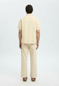 Camicia a maniche corte di colore giallo chiaro con un motivo testurizzato, abbinata a pantaloni beige a gamba larga. Calzature informali di colore marrone.