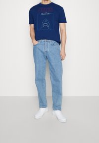 Marine katoenen t-shirt met 'JAWS' opdruk en haaienafbeelding, gecombineerd met lichtblauwe denimjeans en witte sneakers, staand op een neutrale achtergrond.