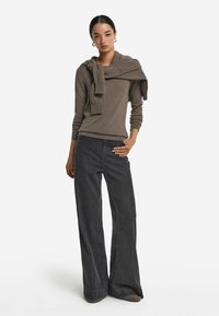 Brązowa, długorękawowa dzianinowa bluzka z zawiązanym swetrem na ramionach, w połączeniu z szerokimi, ciemnymi dżinsami. Charakteryzuje się miękkim materiałem i luźnym krojem.