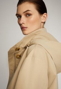 Beige jas met een ritssluiting en bruine knopen, voorzien van een capuchon en een relaxte pasvorm. Soepele textuur, ontworpen voor casual gebruik.