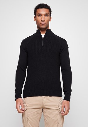 Pull noir en maille côtelée avec col zippé quart et manches raglan, présentant une surface texturée et des poignets ajustés. Fond neutre.