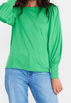 Kobieta ubrana w jasnozieloną, długorękawową bluzkę z bufkami na rękawach oraz jasnoniebieskie dżinsy na tle jednolitego tła.