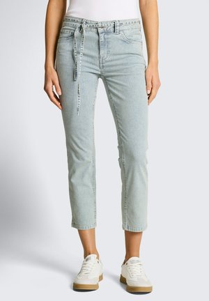 Frau trägt hellblaue und weiße, vertikal gestreifte, gekürzte Hose mit Bindegürtel und weißen Turnschuhen, steht vor einfachem Hintergrund.