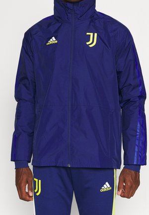 Mężczyzna ubrany w granatową bluzę sportową Adidas i pasujące spodnie z logo Juventusu oraz żółtymi akcentami, stoi ze zrelaksowanymi ramionami.