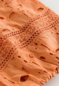 Tissu orange avec bordure en dentelle complexe et motifs découpés brodés sur une surface textile douce et texturée.