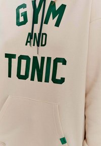 Beige mikina s kapucí a zeleným nápisem "GYM AND TONIC". Obsahuje šňůrkovou kapuci, klokankovou kapsu a malou zelenou výzdobu na dolním lemu.