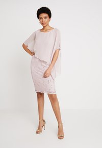 Robe en dentelle rose avec un superposé lâche et doux ; présente un ourlet festonné et de délicates embellissements ; associée à des sandales à talons hauts dorées.