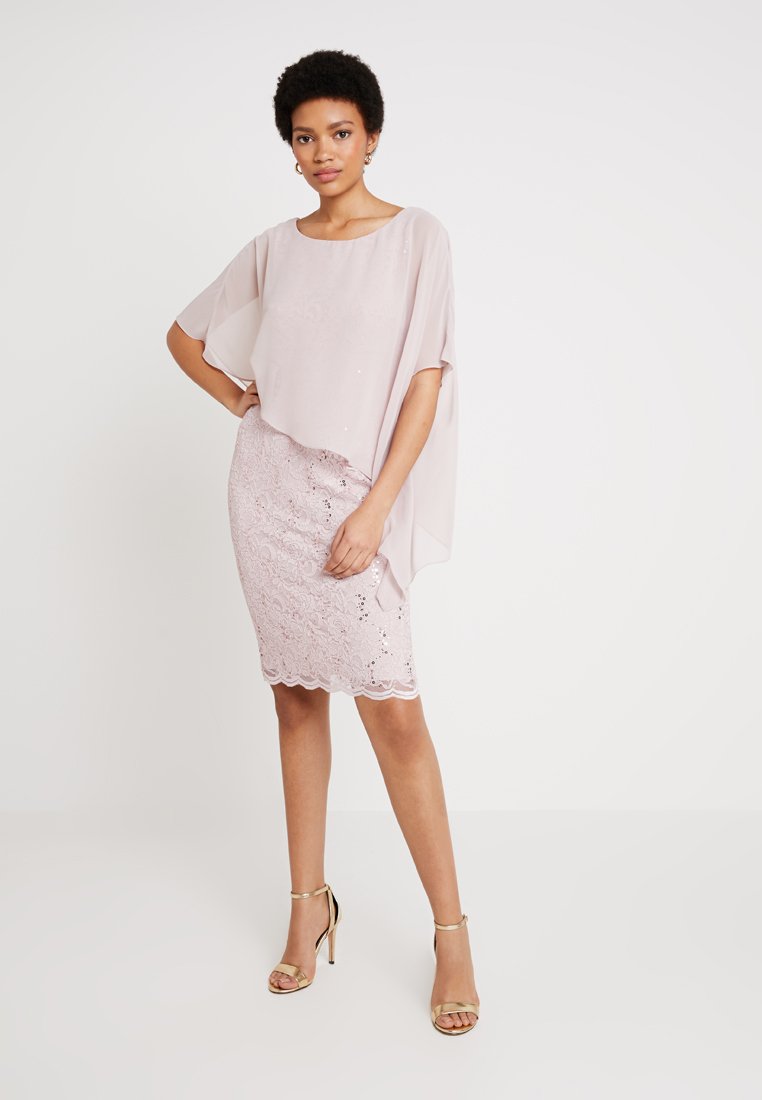 Robe en dentelle rose avec un superposé lâche et doux ; présente un ourlet festonné et de délicates embellissements ; associée à des sandales à talons hauts dorées.