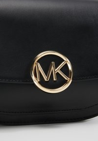 Schwarze Lederhandtasche mit glatter Textur. Merkmale ein auffälliges goldenes Rundlogo mit "MK" in der Mitte. Abgerundet mit sauberem Stich.
