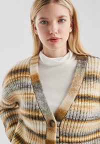 Gestreifter Cardigan mit weitem V-Ausschnitt in Gelb-, Braun- und Grautönen. Strukturiertes Strickmaterial mit Knopfverschluss, über einem weißen Rollkragenpullover getragen.