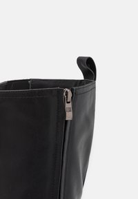 Bolso de cuero negro con una textura suave. Cuenta con un cierre de cremallera y un pequeño asa. El diseño es minimalista y angular.