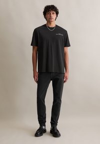 Zwarte katoenen T-shirt met korte mouwen, ronde hals en een klein wit logo accent op de linkerborst, gecombineerd met zwarte jeans en donkere schoenen.