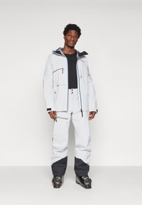 Chaqueta impermeable blanca con detalles en gris y múltiples bolsillos con cremallera, combinada con pantalones a juego y botas de esquí negras.