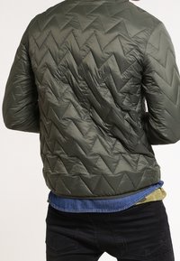 Muž v olivově zelené prošívané bundě se zkříženým stehem, oblečený na modrou košili a černé kalhoty, viděn zezadu.