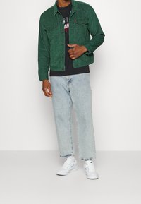 Chaqueta de pana verde con cierre de botones, camiseta negra con estampado, vaqueros azul claro de pierna recta y zapatillas blancas.