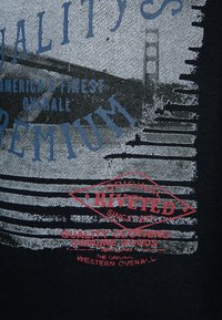 Schwarzer Stoff mit weißem und blauem abgenutztem Text und einem roten Diamant-Logo mit der Aufschrift "ORIGINAL RIVETED". Enthält eine Grafik einer Brücke.