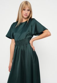 Temno zelena satenasta obleka s kratkimi, razširjenimi rokavi, pasom okrašenim z čipko in svobodnim krilom. Gladka tekstura, eleganten dizajn.