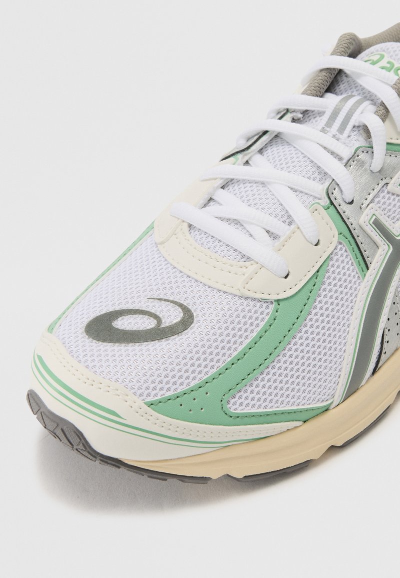 Zapatilla deportiva blanca con parte superior de malla, acentos verdes y plateados, logo redondo, cordones estándar y suela acolchada para mayor comodidad.