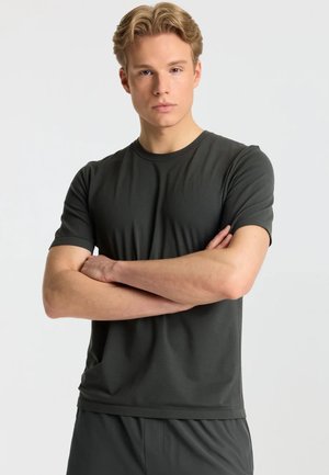 Ung mand med lyst hår står med armene krydset iført en mørkegrå t-shirt med korte ærmer og matchende bukser mod en ensfarvet baggrund.