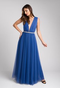 Długa niebieska suknia z tiulu z głębokim dekoltem w kształcie litery V i plisowanym wzorem. Paskiem w srebrnym kolorze w talii. Modelka w czarnych szpilkach.