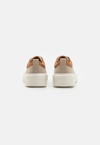 Zapatillas de lona beige con un detalle de cuero marrón en el talón, que cuentan con suela de goma y cordones planos. Textura suave y diseño minimalista.