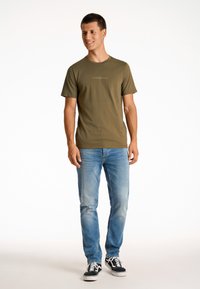 Camiseta de algodón color verde oliva con mangas cortas y cuello redondo, que presenta un sutil logo estampado en un color claro en el pecho. Combinada con jeans azules desgastados y zapatillas negras.