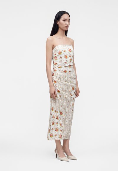 Femme aux longs cheveux noirs portant une robe blanche bustier à imprimé floral rose et jaune, debout avec une expression neutre et des talons hauts beiges.