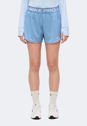 Personne portant un short Nike Pro bleu clair, un haut blanc à manches longues, des chaussettes blanches et des baskets Nike blanches, debout sur un fond uni.