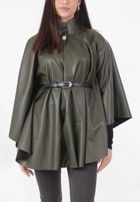 Olijfgroene cape van imitatieleer met een hoge kraag, vastgemaakt met een zwarte riem. Heeft een glanzende zilveren knop bij de kraag.