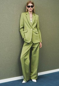 Op maat gemaakt groen blazer en wijde broek in een soepele stof, gecombineerd met witte puntige schoenen en grote zwarte zonnebril.