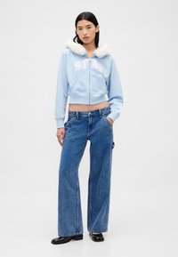 Felpa corta azzurro chiaro con cappuccio foderato in pelliccia sintetica bianca e scritta "STAY". Abbinata a jeans blu a gamba larga e stivali neri. Design minimalista.