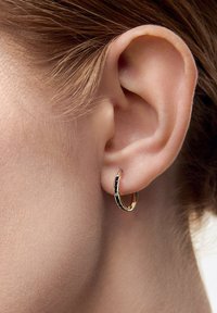 Złoty kolczyk w kształcie koła z akcentem z czarnej emali. Okrągły design, gładkie wykończenie i mały detal zapięcia. Noszony w przekutym uchu.