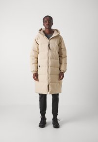 Manteau rembourré beige avec capuche, fermeture éclair sur le devant et bandes élastiques aux poignets. Présente un design jusqu'aux genoux et une texture matelassée.