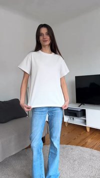 Femme aux longs cheveux foncés porte un t-shirt blanc et un jean bleu, debout dans un salon avec canapé, télévision et sol en bois.