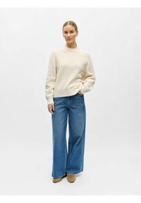 Kermainen neulepaita, jossa on korkea kaulus ja resorit hihansuissa, yhdistetty rentoon tyyliin leveälahkeisten sinisten farkkujen kanssa.