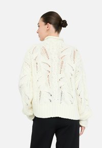 Donna con un chignon intrecciato che indossa un maglione bianco a trecce con motivi a foglia e pantaloni neri, vista di spalle su uno sfondo bianco.