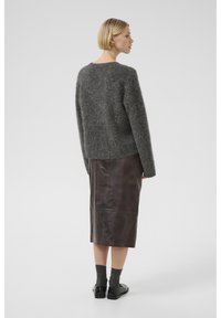 Pull en maille grise et duveteux à longues manches, associé à une jupe midi en cuir marron. Le modèle porte des chaussettes grises et des chaussures noires.