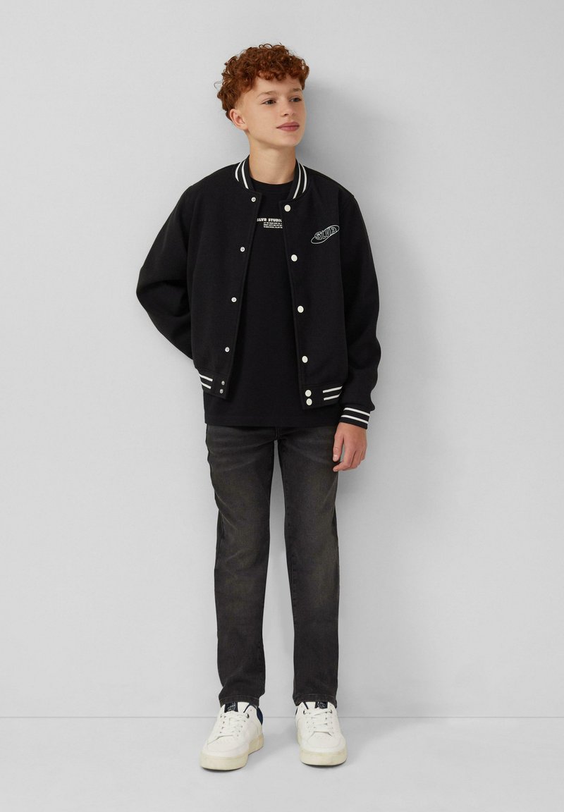 Zwarte varsityjack met witte gestreepte manchetten en kraag, gedragen over een zwart t-shirt, in combinatie met donkere jeans en witte sneakers.