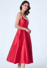 Robe A-line en satin rouge avec un col ornée de perles, dotée de fines bretelles, d'un corsage ajusté et d'une jupe fluide atteignant le mi-mollet.