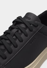 Μαύρο δερμάτινο αθλητικό παπούτσι με υφή στη φτέρνα, μαύρα κορδόνια και ανάγλυφο λογότυπο "BOSS" στη γλώσσα και την πλευρά, με ορατή μπεζ καουτσούκ σόλα.