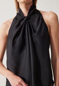 Top halter nero realizzato in tessuto liscio, con un design annodato al collo e morbide drappeggiature. Nessun hardware o decorazione visibile.