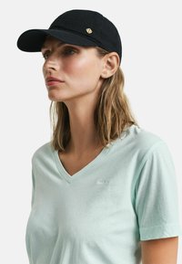 Sorte kasket med en buet skygge og et guldemblem på siden, båret med en lys mintgrøn v-hals t-shirt. Glat bomuldsstruktur.