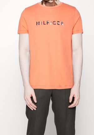Personne portant un t-shirt orange à manches courtes avec le texte "HILFIGER" sur la poitrine et un pantalon foncé, debout devant un fond uni.